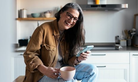 Middle age woman having coffee break and using phone in the kitchen at home during daytime. She is looking at camera and smiling with positive attitude while texting message on cellphone.