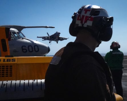 A sailor with their back to the camera watches as a fighter jet comes in to land on an aircraft carrier