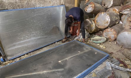 A man bends over a large metal panel. Cylinders for holding water are stacked behind him