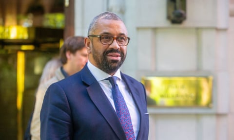 Midshot of James Cleverley in blue suit and tie outside building with large brass plaque on wall