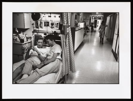 John lying on a hospital bed, chatting with another man who is lying in the same bed. From ‘The Ward’ by Gideon Mendel, 1993