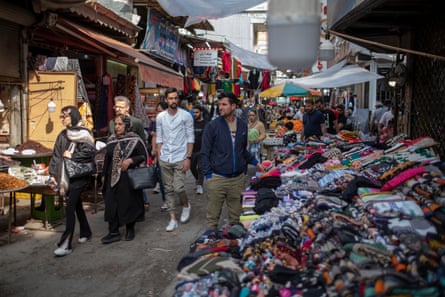 A stallholder stands next to tables piled with olourful clothing, while around him can be seen dried fruit stalls and other places selling produce and goods.