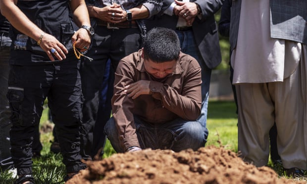 A man kneels down, crying, in a cemetery surrounded by other mourners.