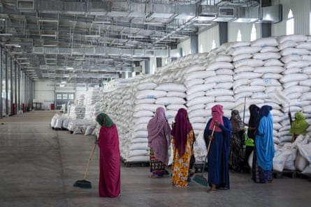 Women stand in a warehouse next to sacks of food aid, piled high.