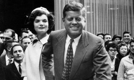 A black-and-white photo of a smiling white man in a suit facing the camera, reaching behind to hold the man of a serene-looking white man with dark curled hair, a light-colored hat and overcoat, with dozens of people behind them.