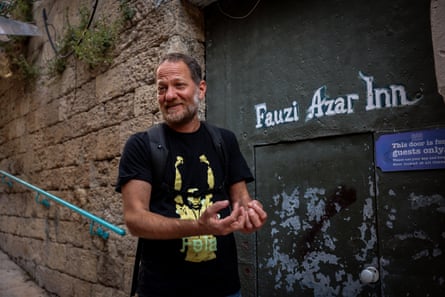 A middle-aged white man in a T-shirt with a picture of Fela Kuti stands outside a metal door set into an old stone wall in an alleyway