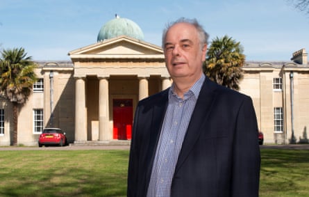 Prof Wyn Evans outside the Institute of Astronomy, which is a stone building with a dome and classical-style pillars and portico at its entrance. He appears to be in his 60s with short grey hair. He wears a blue and white shirt with small checks under a dark blue jacket.