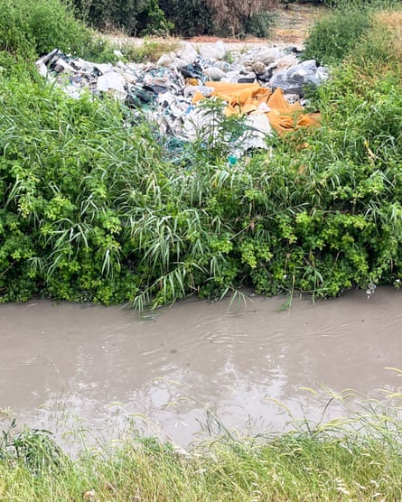 A pile of plastic rubbish amid greenery by a narrow conduit full of dirty grey-brown water