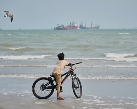 A boy with his bicycle watches a gull fly over the sea, ships on the horizon