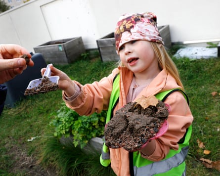 A child wearing hi-vis holds a small plastic toy shovel with part of a dirt cake on it. She holds the rest of the dirt cake in her other hand