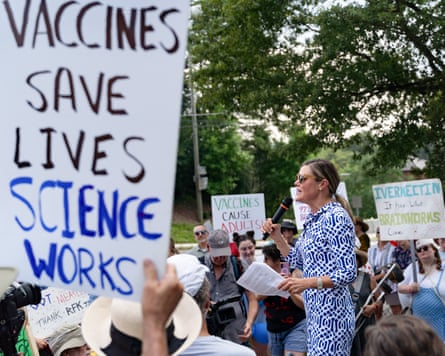 A woman holds a microphone and speaks to a crowd holding protest signs.