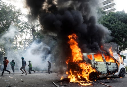 Smoke billows from a burning car as people run past
