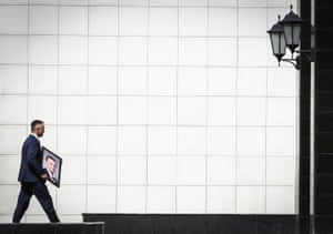 A man carries a framed photograph towards a black doorway against a backdrop of a white tiled wall