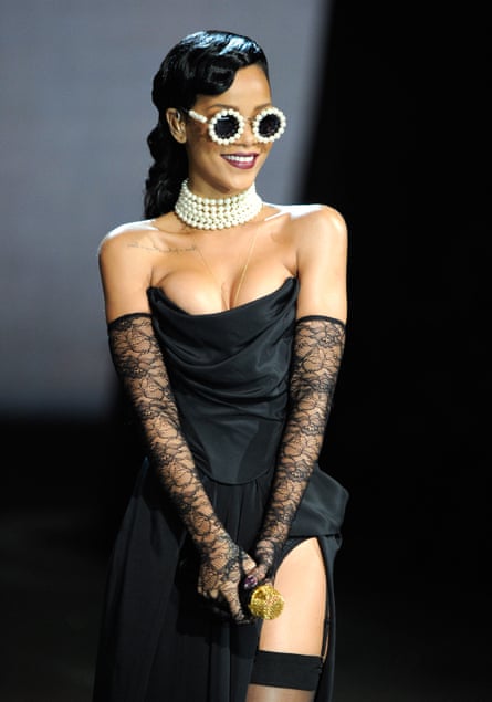 A woman with dark shiny hair wearing a revealing short black satin sleeveless dress and long lacy gloves and sunglasses with pearls around them