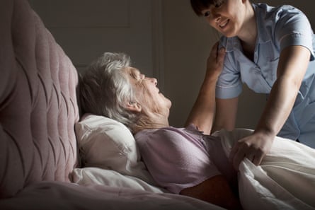 An elderly woman is tucked into bed at home by a care assistant
