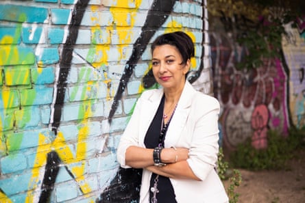 she is in her mid-50s with dark hair and red lipstick; she wears a black top and white jacket, and is standing with her arms folded in front of a brick wall with colourful graffiti.