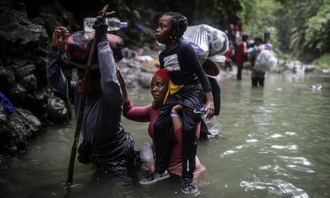 a woman wades through waist-high water while carrying a child on her shoulder next to another woman carrying a bag on her head