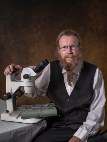 A man rests his hand on a microscope. A book lies on the base of the microscope