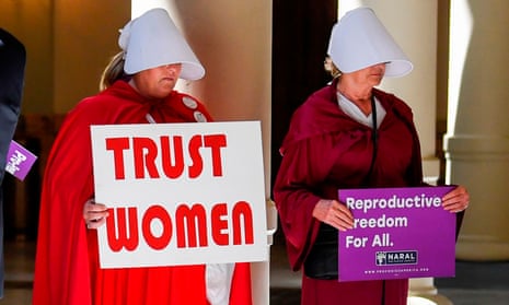 Women in Handmaid's Tale costumes with signs reading 'Trust women' and 'Reproductive freedom for all'.