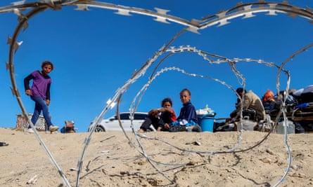 Palestinian children sit on sand with barbed wire in front of them among their belongings