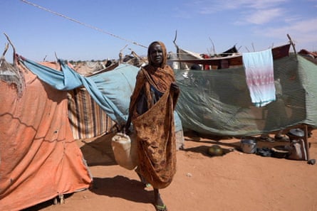 A woman carries a water container at a camp for displaced people: she wears a patterned brown scarf around her head and body and steps between makeshift tents and shelters made from sticks and orange, blue and green cloth, on reddish-brown earth.