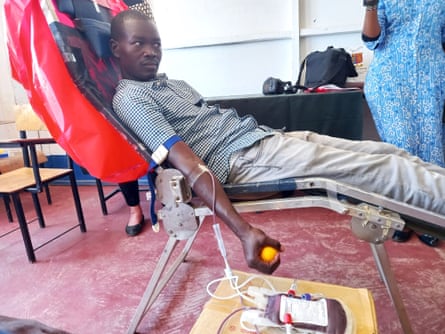 A young man sits on a reclining chair. He has a cannula in his arms and blood flows from it into a bago n a low table beside him. He is holding a ball to keep the pressure up.