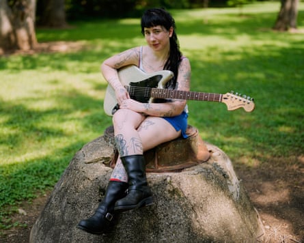 Karly Hartzman holding a guitar while sitting on a concrete mound with a metal top, with grass and trees in the background.