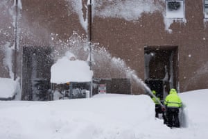 Snow sprays from a clearer as a person cuts a gap through the snow outside the door to a building