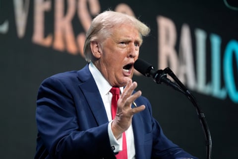An older white man in a blue suit, white shirt and red tie, speaking and gesturing on a stage.