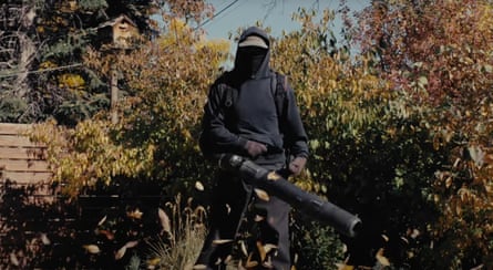 A man dressed all in black holds a leaf blower in front of a garden full of autumn trees, red and green coloured.