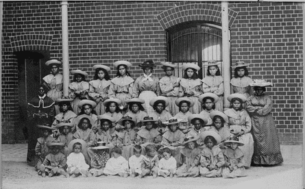 A black and white image of a group of Aboriginal girls wearing dresses and hats lined up in four rows
