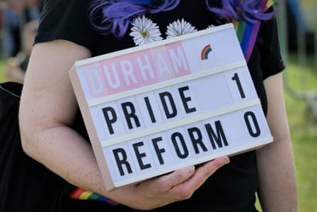 A person holds a board which has the word Durham followed by a rainbow. Underneath is says Pride 1, Reform 0