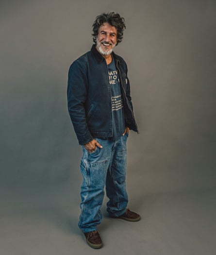 Lorenzo Tondo standing with his hands in his pockets, smiling at the camera, wearing a blue T-shirt with beige writing on, an unzipped blue workwear jacket, baggy blue jeans and brown trainers, against a grey background
