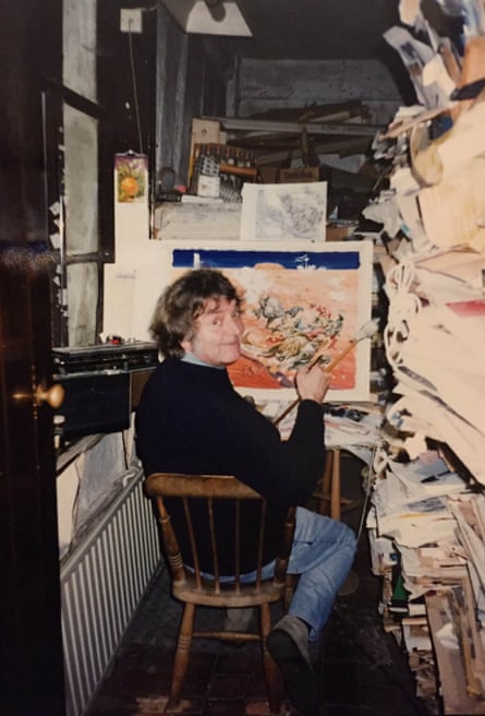 Josh Kirby smiles over his shoulder while seated in front of a painting with a paint brush in his hand. The room is narrow and cramped with papers on one side