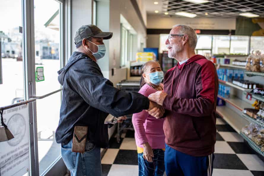 A man wearing a protective face mask greets another man inside a grocery store. A woman in a mask watches the interaction.