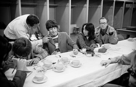The Beatles, Baez and Gleason sitting at a long table covered with a white tablecloth, drinking tea while McCartney talks into a microphone held by Mitchell
