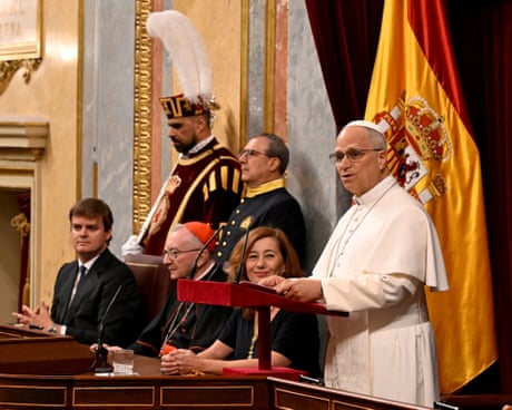 Pope Leo speaks at a red podium, with officials and a Spanish flag behind