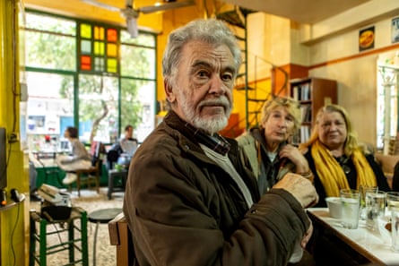 Stelios Goutis sits at the bar with two women to his side and glasses in front of him. He is in his early 80s with white hair and a white beard. There is stained glass in the windows behind him and people are sitting at tables.