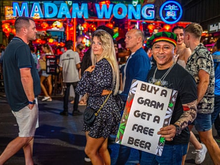 A man holds a sign saying: ‘Buy a gram get a free beer’ as  tourists walk behind him and a neon blue shop sign says: ‘Madam Wong’