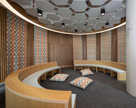 Three cushion lie on the floor surrounded by circular wooden benches inside a circular room with geometric patterned walls breaking up wooden panelling