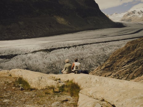 Two people sit on a rocky ledge above the glacier with snowy peaks in the background