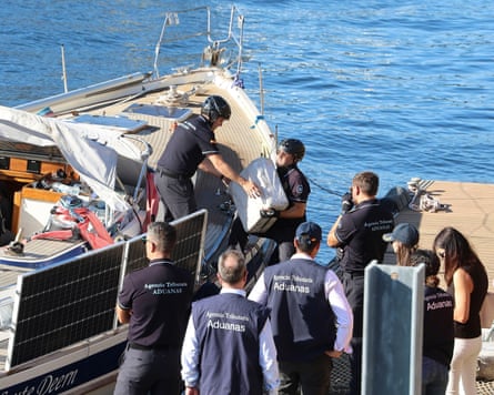 Men in shirts marked Aduanas (Spanish for customs) carry large packages of a sailing boat at a quay