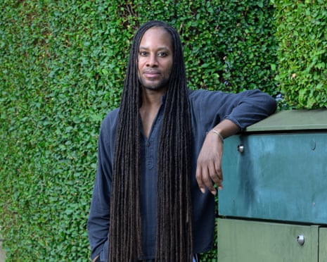 Man with long dreadlocks leans against a cabinet in front of a hedge