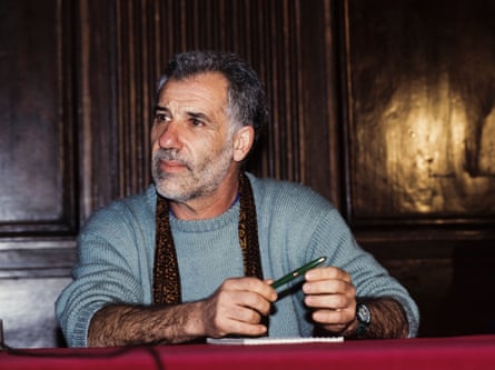 Renato Curcio, middle-aged with a short grey beard, sits at a desk, holding a fountain pen.