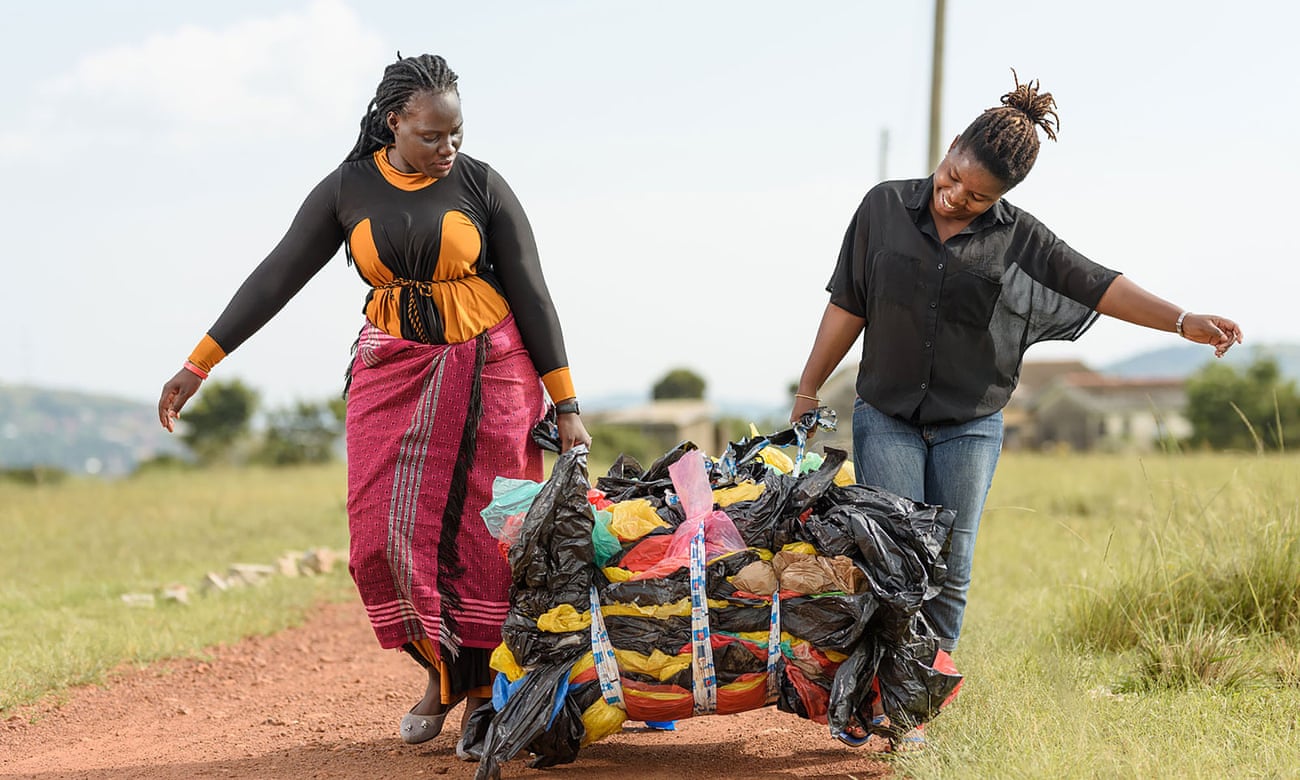 Two Ugandan women carry a bundle of waste plastic along a red dirt road.