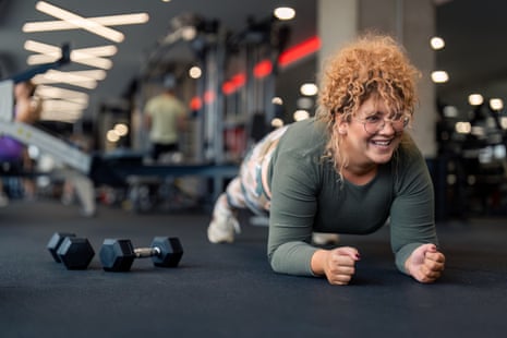 Determined smiling young woman with curly hair in plank positionDetermined smiling young woman gym fitness client with curly hair in plank position doing exercises for strong core abdominal muscles having dumbbell weight set next to her focused on fitness goal at gym.