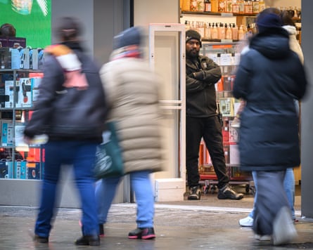Shoppers walk past a store with a security guard standing in the doorway.