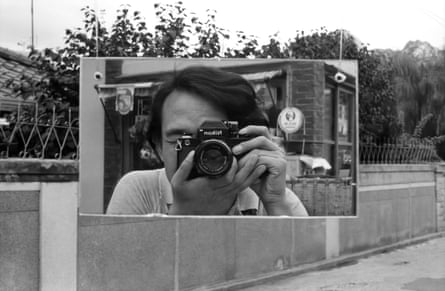 A black-and-white photo of an image within an image. The center is a young man with long black bangs holding a camera to his face, as if taking his picture in a mirror. This sits in the middle of a pedestrian-looking cement walkway bordered by trees.