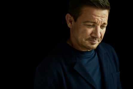 Portrait of Jeremy Renner slightly smiling while looking down. He is wearing a dark T-shirt and jacket and shot against a black background