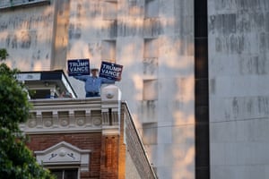 A man standing on the corner of a rooftop holds up signs reading 'Trump Vance' while people out of shot take part in a protest
